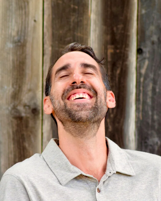 Sarkis Love laughing with his eyes closed, in front of a weathered wooden wall